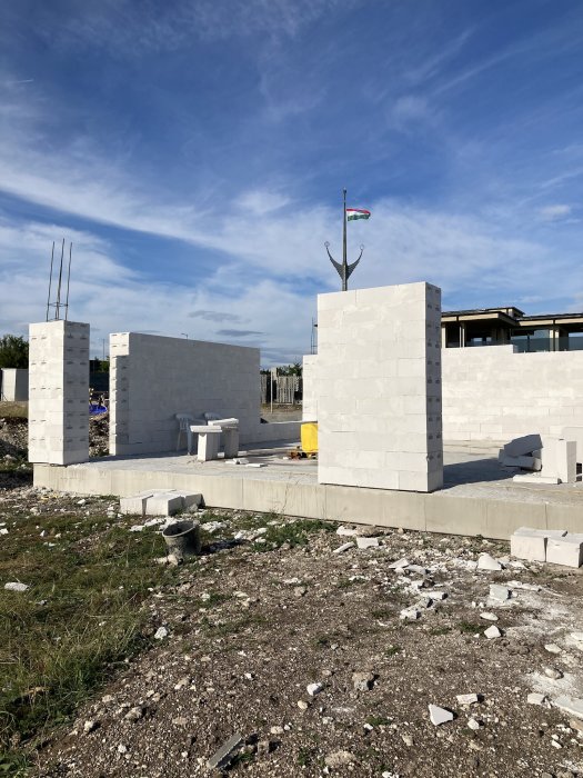 Byggarbetsplats med pågående konstruktion, betongblock, flagga på stolpe, blå himmel, och byggmaterial på marken.