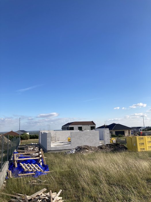 Byggarbetsplats med pågående husbyggnation under klarblå himmel, material och gräsmark i förgrunden.