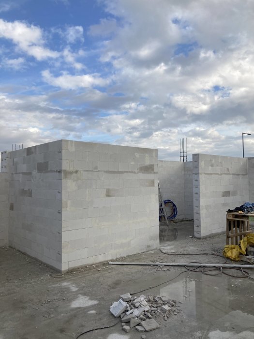 Byggarbetsplats med oavslutade betongväggar under en molnig himmel, byggmaterial och verktyg på marken.