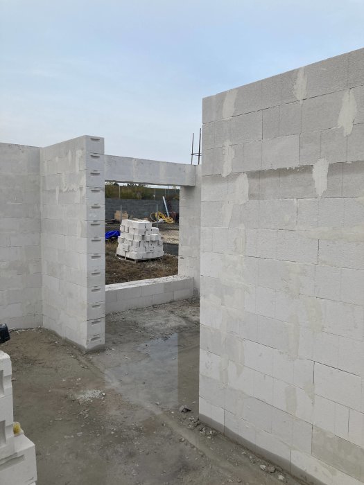 Byggarbetsplats med oavslutade vita väggar av betongblock, vattenpussar på marken, byggmaterial i bakgrunden.