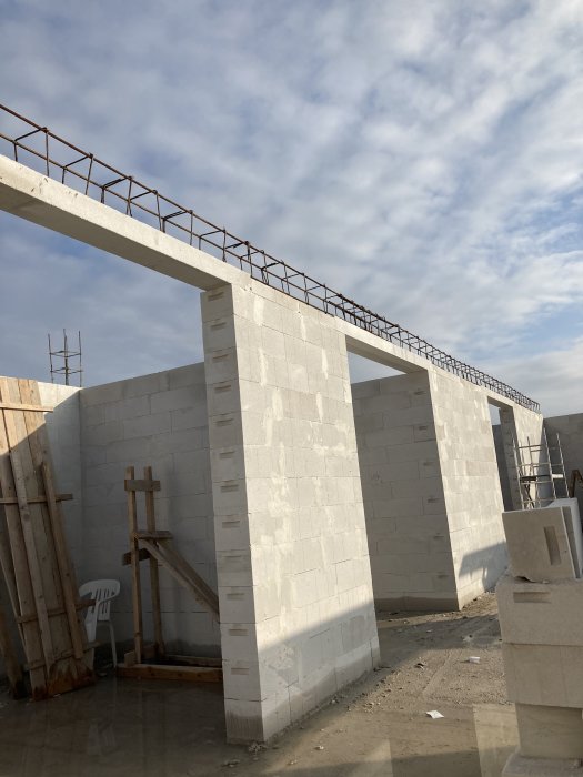 Byggarbetsplats med betongväggar och balk, blå himmel, ställningar, byggmaterial på marken.