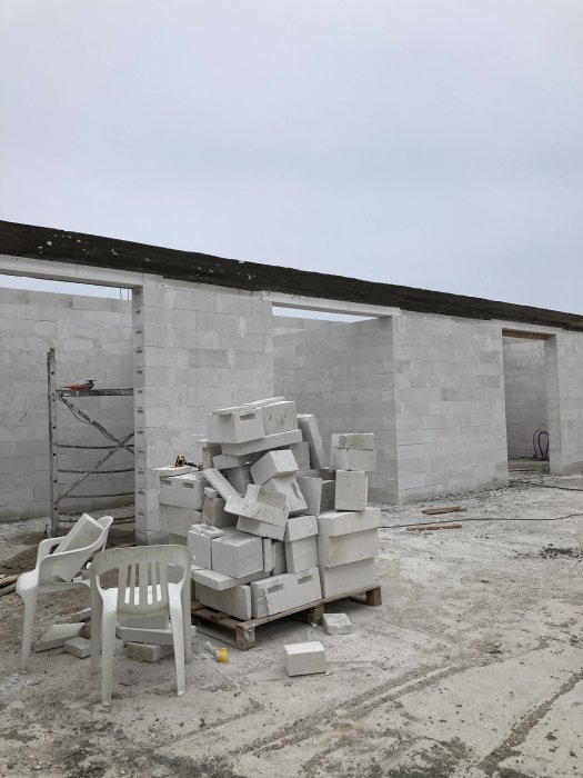 Byggarbetsplats med vita cellbetongblock, tomma plaststolar och byggställning under en grå himmel.