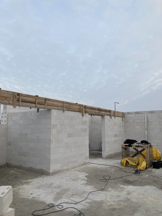 Byggarbetsplats med oavslutade betongväggar, träbjälkar, byggmaterial och verktyg under tidig morgon eller skymning.