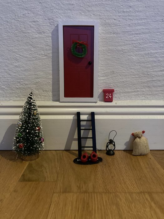 Miniatyr julscen med röd dörr, krans, gran, stege, skor och lykta.