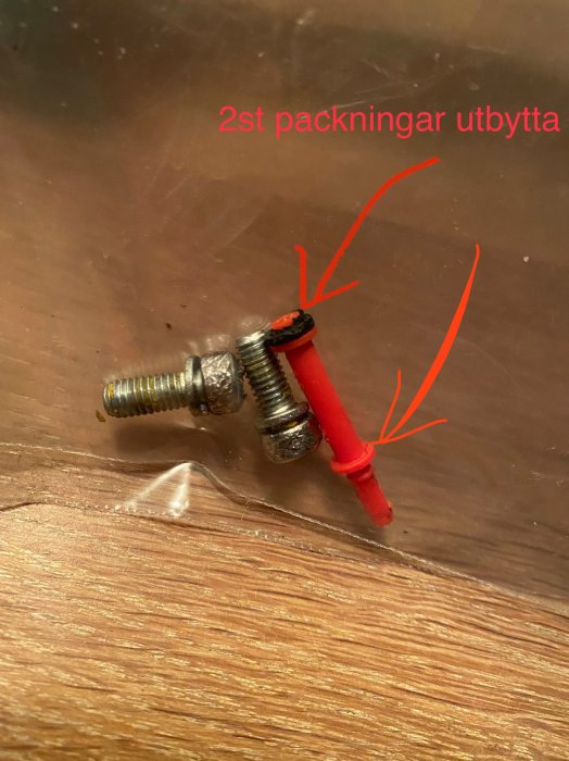 Bult med två utbytta packningar, röda pilar pekar, textnotering, reflektion på träyta.