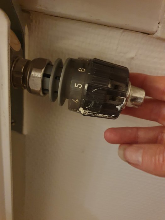 Termostat på radiator, inställd på nivå 4, hand justerar den, vit vägg i bakgrunden.