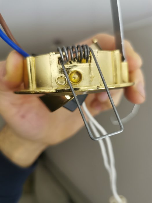 Hand håller en termostatkomponent med fjädrar, skruvar och elektriska ledningar. Fokus på objektet, bakgrund suddig.