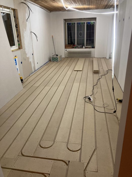 Rum under renovering, tomma väggar, fönster, golvvärmeinstallation innan slutgolv läggs, verktyg och byggmaterial synliga.