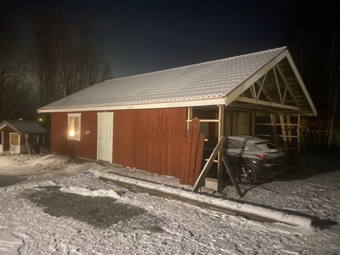 Röd byggnad med snötäckt tak, bilparkering, träd och snöig mark på natten.