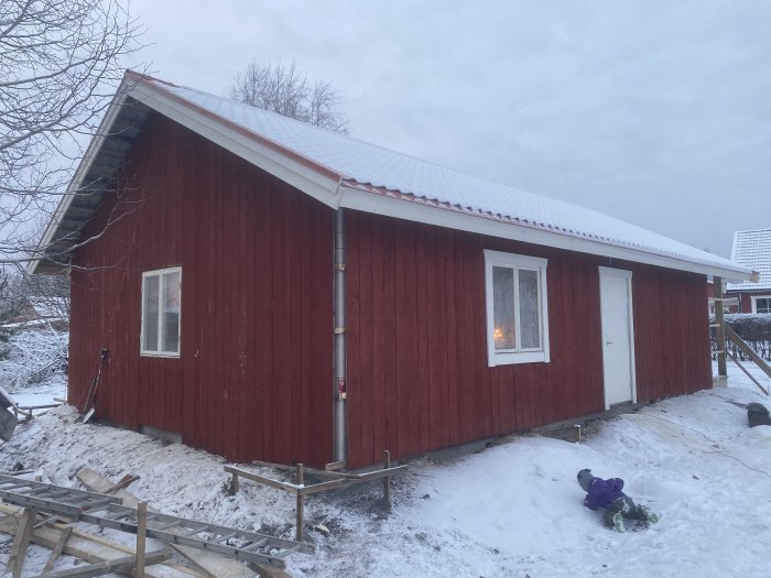 Rött hus i snöigt landskap, byggnadsarbete pågår, barnleksaker framför huset, vintertid.