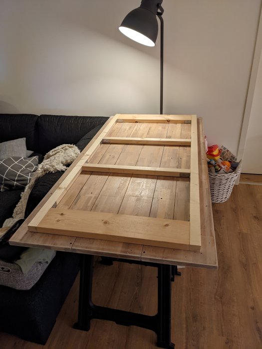 Ett träbord med uppfällda ben framför en soffa, golvstående lampa till vänster, korg med leksaker.