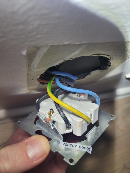 Elektrisk vägguttag under installation, ledningar synliga, skadad gipsvägg, hand håller uttaget.