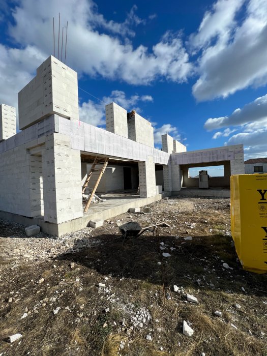 Ett pågående husbygge med vita murblock, armeringsjärn, byggställningar och en blå himmel med moln.