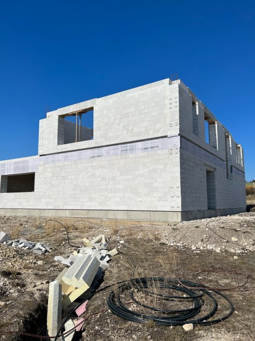 Ett ofärdigt hus av ljusa betongblock, byggarbetsplats med rör och spill, klarblå himmel.