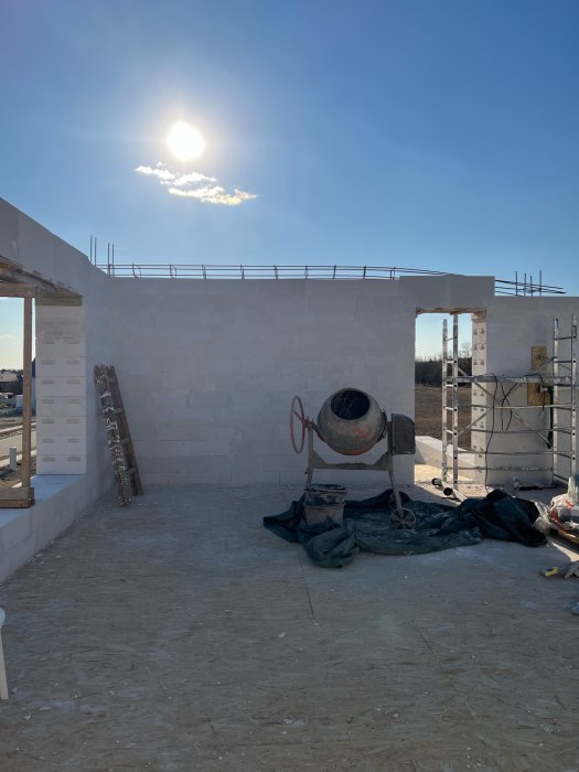 En byggarbetsplats med tegelväggar under en klar himmel, betongblandare i mitten, stegar och byggnadsställningar.