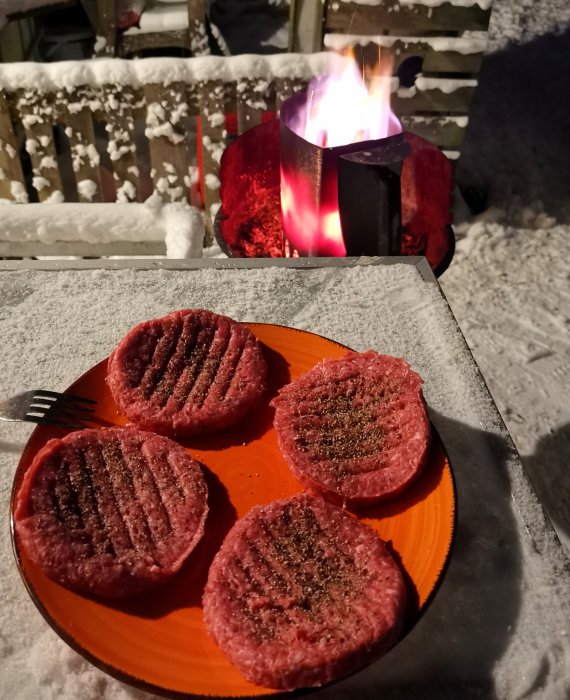 Fyra kryddade hamburgare på orange tallrik, ute, snöig omgivning, bakgrund med brinnande eldpall.