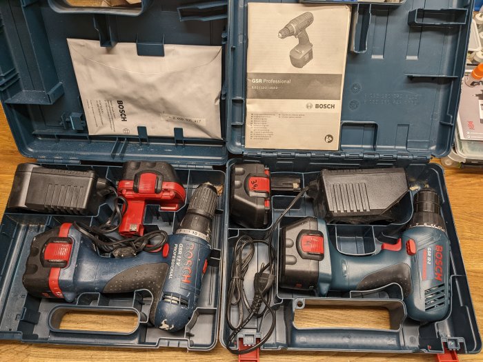 Två Bosch borrskruvdragare med batterier och laddare i öppen verktygslåda, bruksanvisningar, använda, arbetsverktyg.