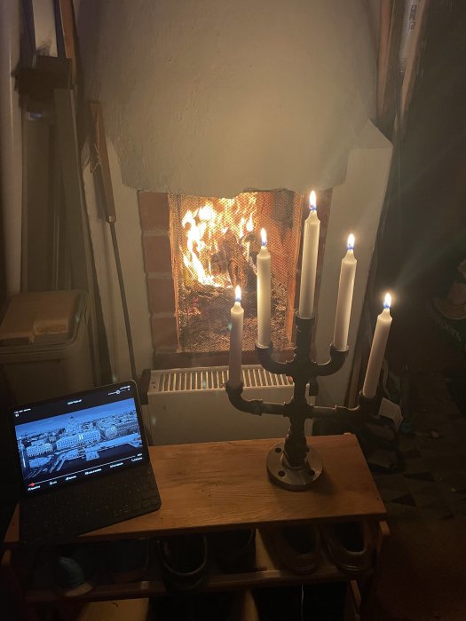 Öppen spis, flammor, tända ljus i ljusstake, laptop med bildskärm, mysig atmosfär, inomhus, kvällstid.