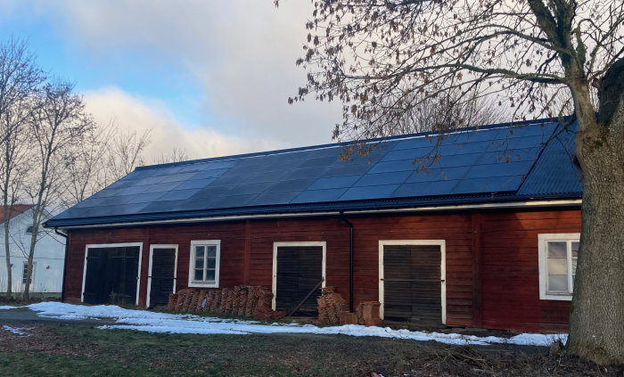 Lång röd träbyggnad med solpaneler på taket, snöfläckar på marken, trädstam i förgrunden.