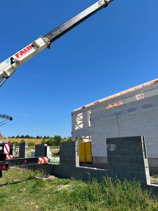 Byggplats med kran och pågående konstruktion av betongblocksvägg under klarblå himmel.