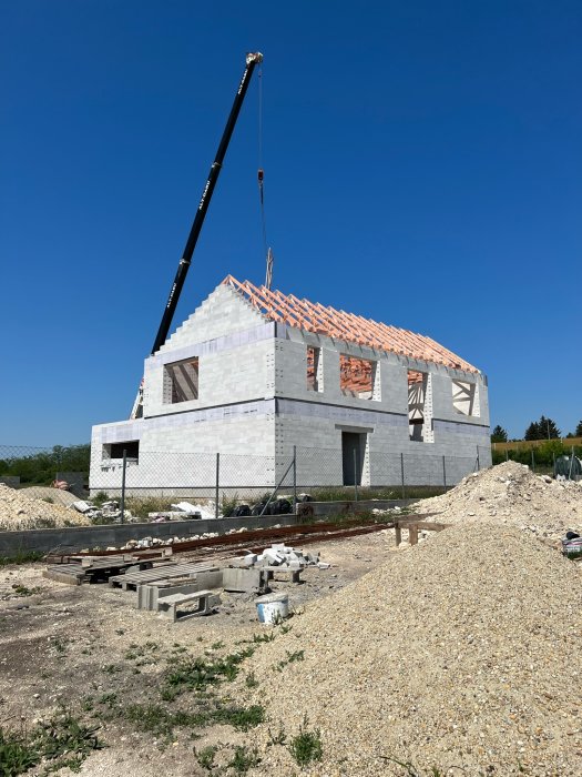 Hus under konstruktion med kran, tegeltak, byggnadsmaterial och blå himmel.