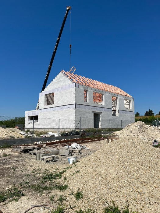 Ett hus under konstruktion med stora vita block, kran, grus och träbjälkar på taket.