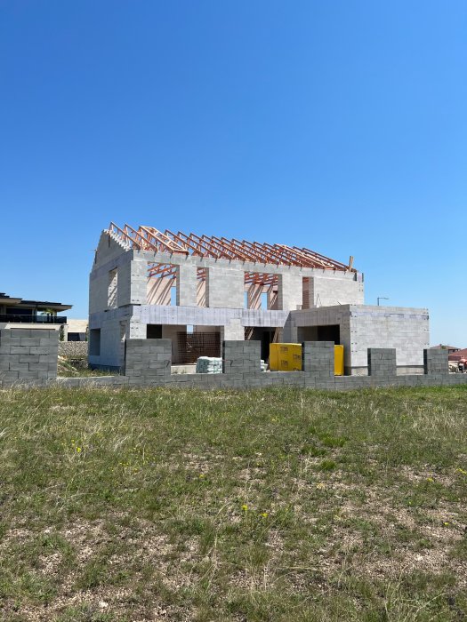 Tvåvåningshus under konstruktion, blå himmel, träbjälkar, grå betongblock, gult byggavfall i förgrunden.