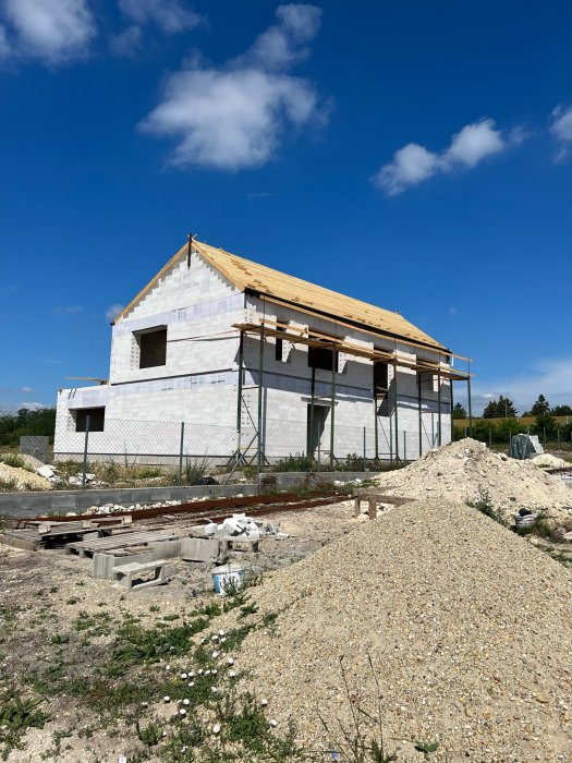 Hus under konstruktion, fasad av skumbetongblock, träbjälklag, byggnadsställningar, blå himmel, byggmaterial på marken.