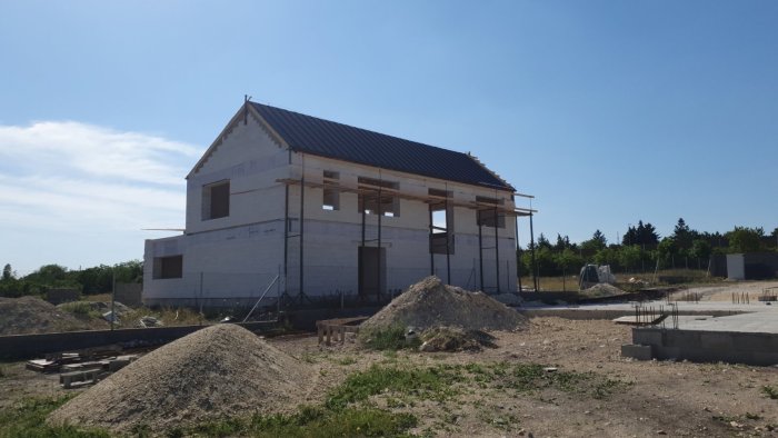 Hus under konstruktion, blå himmel, byggnadsställningar, grushögar, obebyggd tomt, byggmaterial på marken.