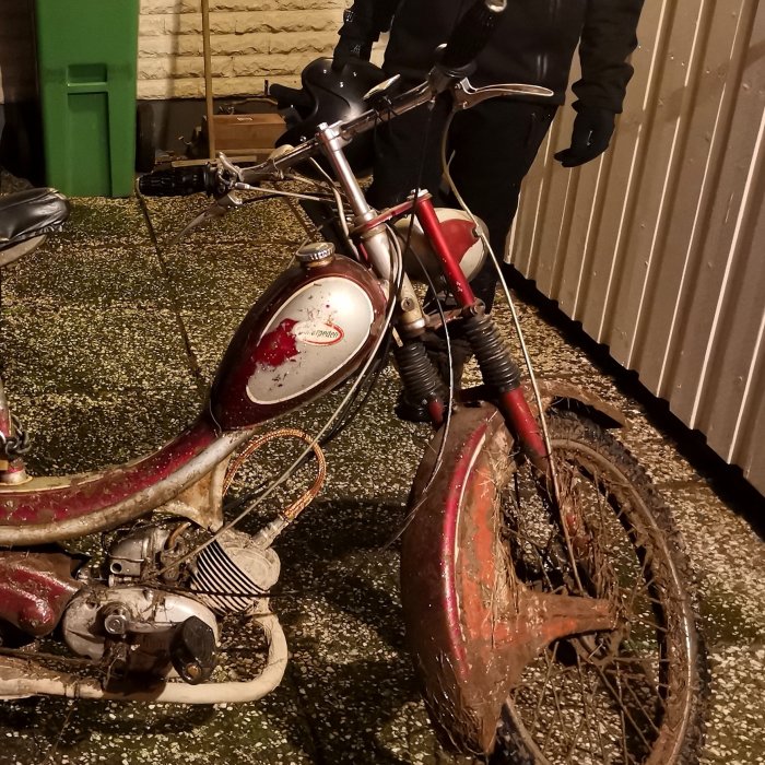 Gammal, rostig moped står utomhus på kvällen med person i bakgrunden.