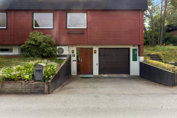 Ett rött radhus med garage, entrédörr, postlåda och luftvärmepump. Vegetation och skog i bakgrunden.