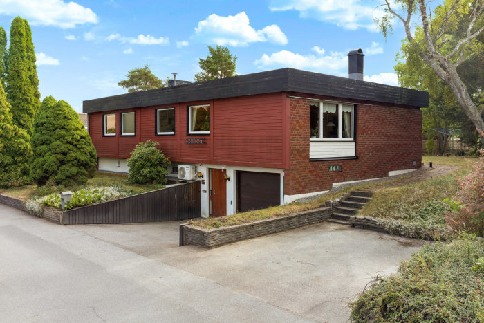 Ett rött tegelhus med garage, grönskande trädgård, blå himmel och lummig omgivning.