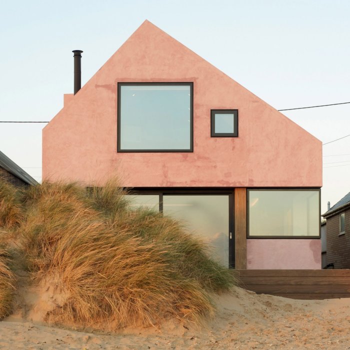 Modernt rosa hus med asymmetriska fönster, skorsten, sanddyner i förgrunden under kvällshimmel.