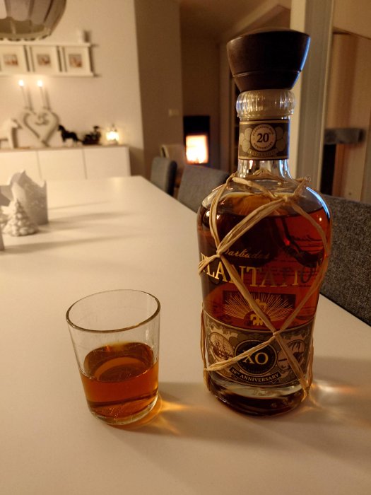 En flaska mörk rom med etiketten "20th Anniversary" och ett glas med rom, mysig inomhusmiljö i bakgrunden.