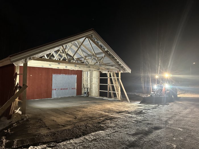 Byggarbetsplats på natten, ofärdigt trähus, traktor med strålkastare, snö på mark, konstruktion av byggnad under ljusa lampor.