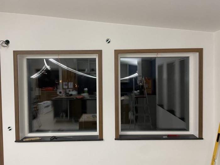 Två fönster i en vit vägg, reflektioner syns, inomhusmiljö, kabeldragning pågår, ett stegpall och verktyg synliga.