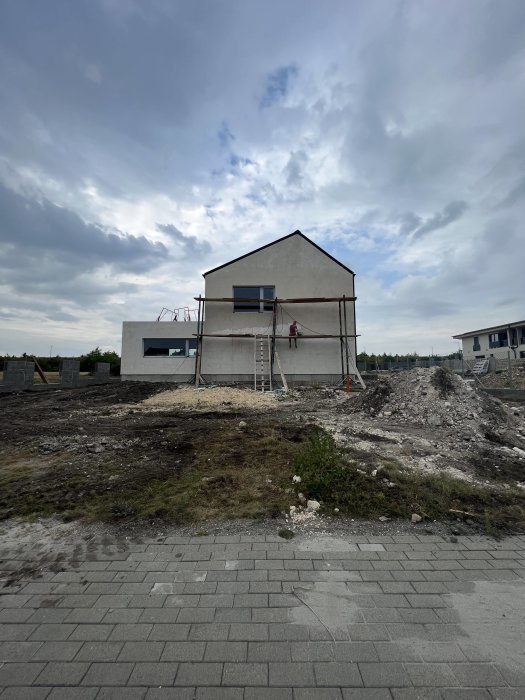 Ett halvfärdigt hus med ställning och stege under molnig himmel. Obyggt landskap med grus och vegetation.