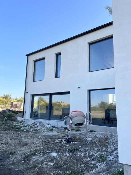 Nybyggt hus, oavslutad konstruktion, byggnadsavfall, cementblandare, hund, stora fönster, klar himmel.