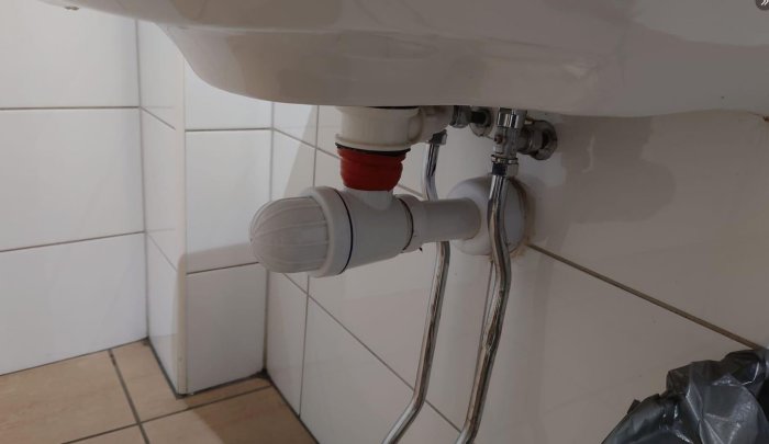 Tvättställ med synliga rörledningar och vattenlås mot kakelklädd vägg, soppåse i förgrunden, sanitära installationer.