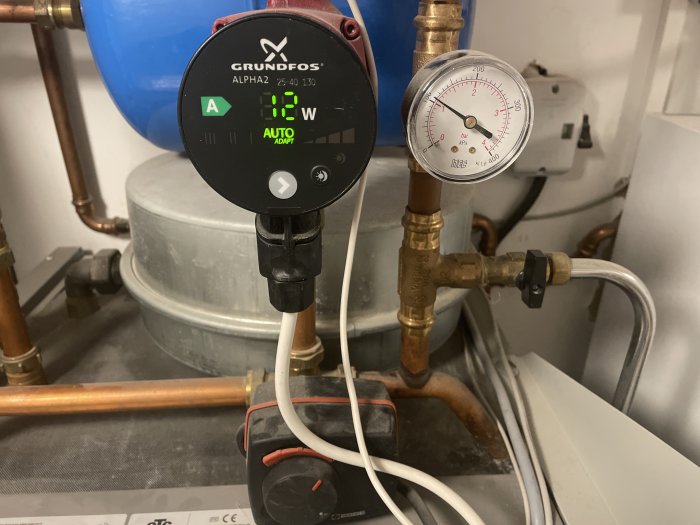 Cirkulationspump och manometer i ett värmesystem, rör, elanslutningar, och en expansionskärl i bakgrunden.