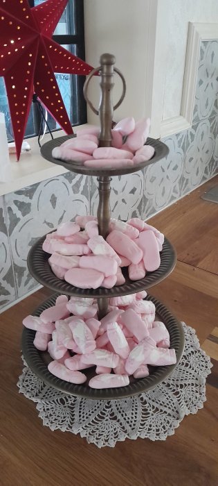 Trefaldig etagere med rosa skumgodis, röd julstjärna i bakgrunden, vita spetsdukar, inomhusmiljö.