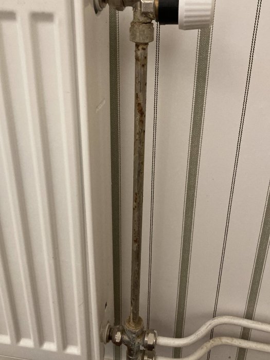 Radiatorkoppling med rostigt rör, termostatventil och vitmålade väggar med tapet.