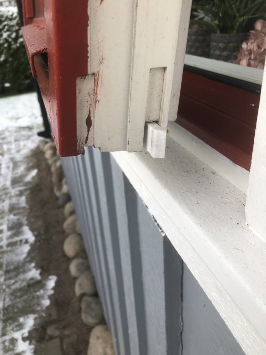 Fönsterlucka på hus med vit skjutbar del, röd karm, snöig trädgård i bakgrunden.