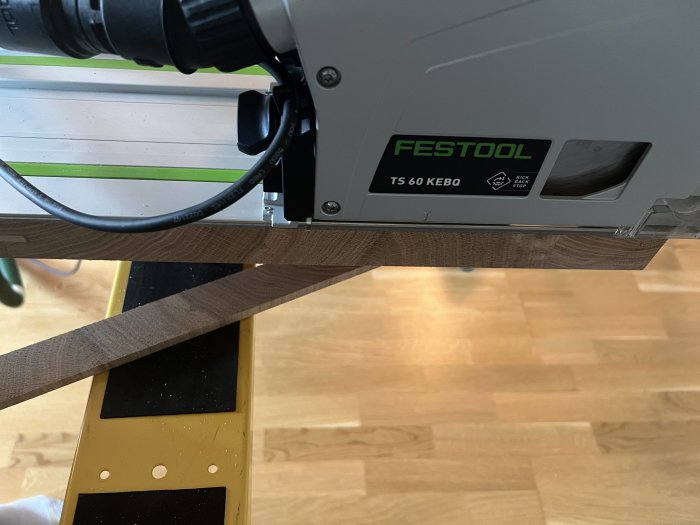 Elektrisk sticksåg Festool TS 60 monterad på skena, sågar träplanka, över gul verktygsbänk, trägolv i bakgrunden.