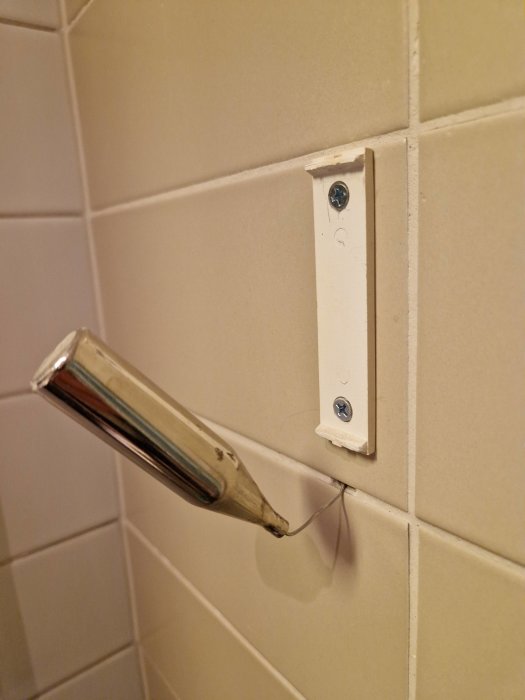 Handtaget till en duschdörr lossnat, hänger löst på en metallskruv, vit kakelvägg i bakgrunden.
