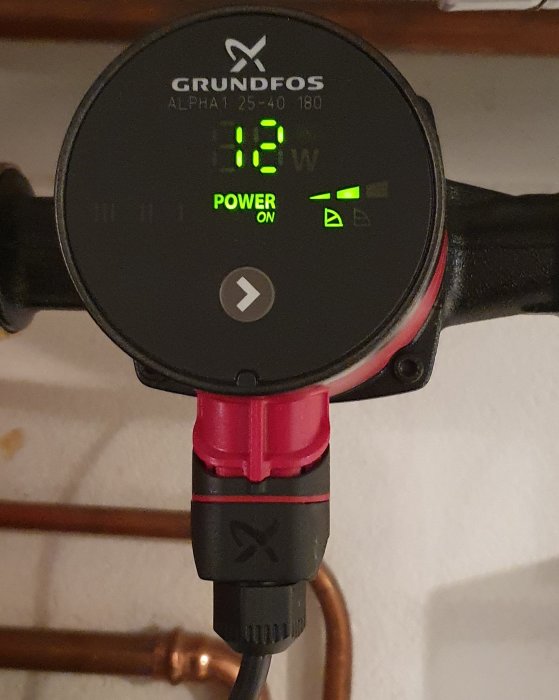 Grundfos cirkulationspump med display, visar 12W effektförbrukning, röda rörkopplingar, svart el-kabel, power on.