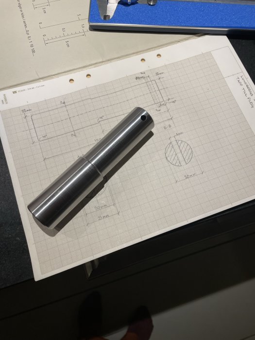 Metallkomponent ligger på teknisk ritning med mått, ingenjörskoncept, designprocess, mätplattform och ritningsutrustning.