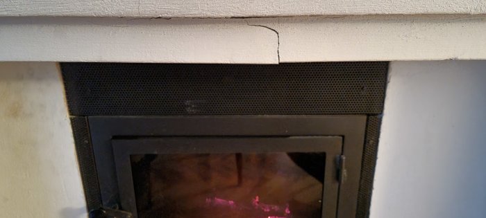 Öppen spis med spricka i väggen ovanför, svart skyddsnät, inredningsdetalj, eld syns inuti.