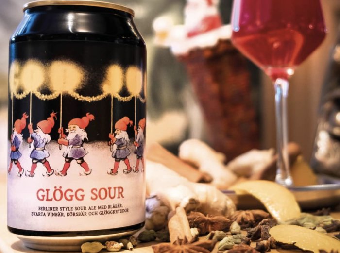 Burk med "Glögg Sour", tecknade tomtar, vinrött innehåll i glas, kryddor och fruktskivor i förgrunden.