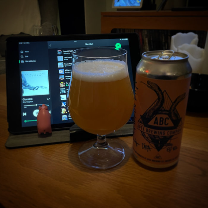 Ett glas öl, en ölburk och en surfplatta som visar en spellista, allt på ett bord.